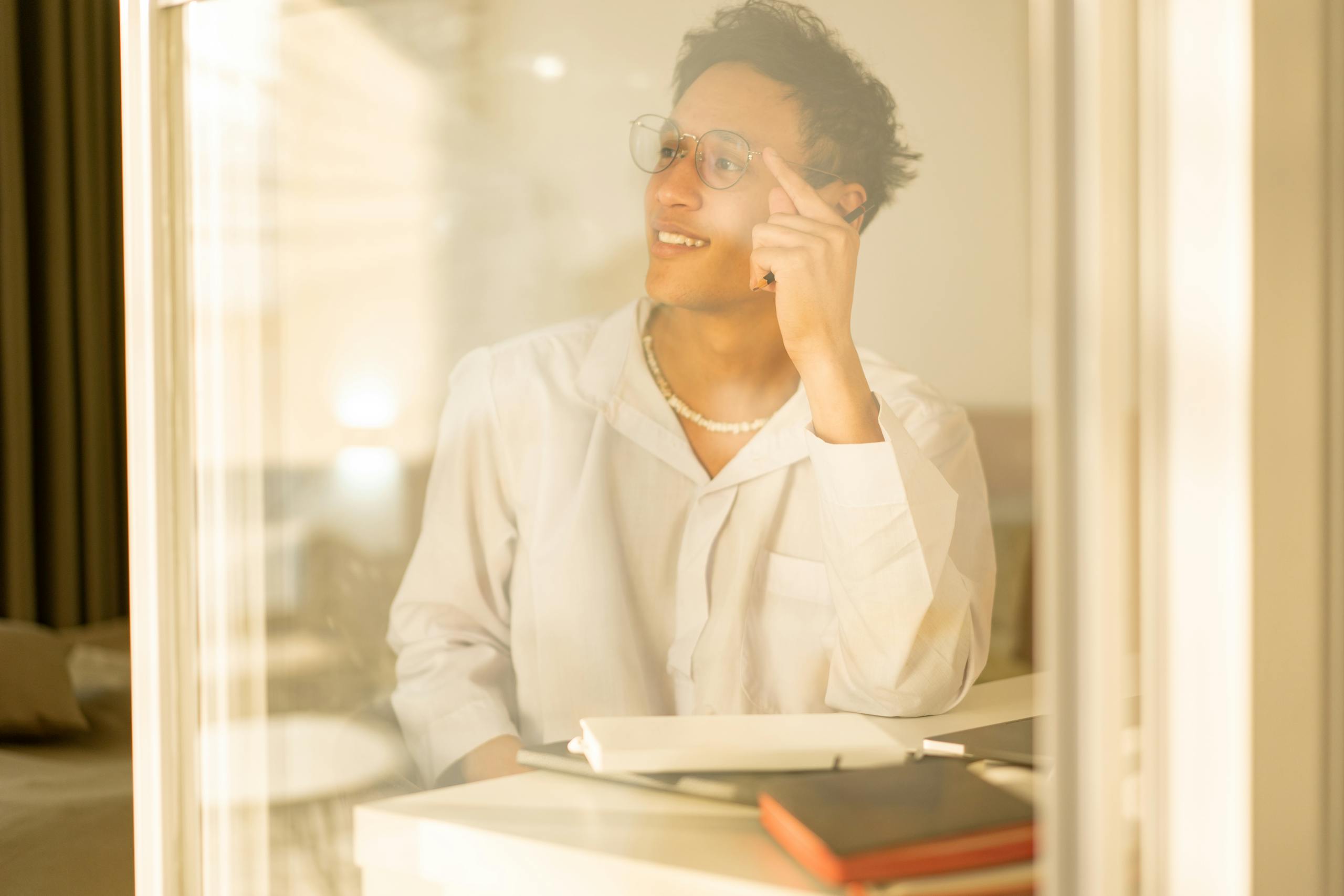 Man with glasses sitting indoors, pensive and relaxed. Clearly reflecting through a window with warm lighting.