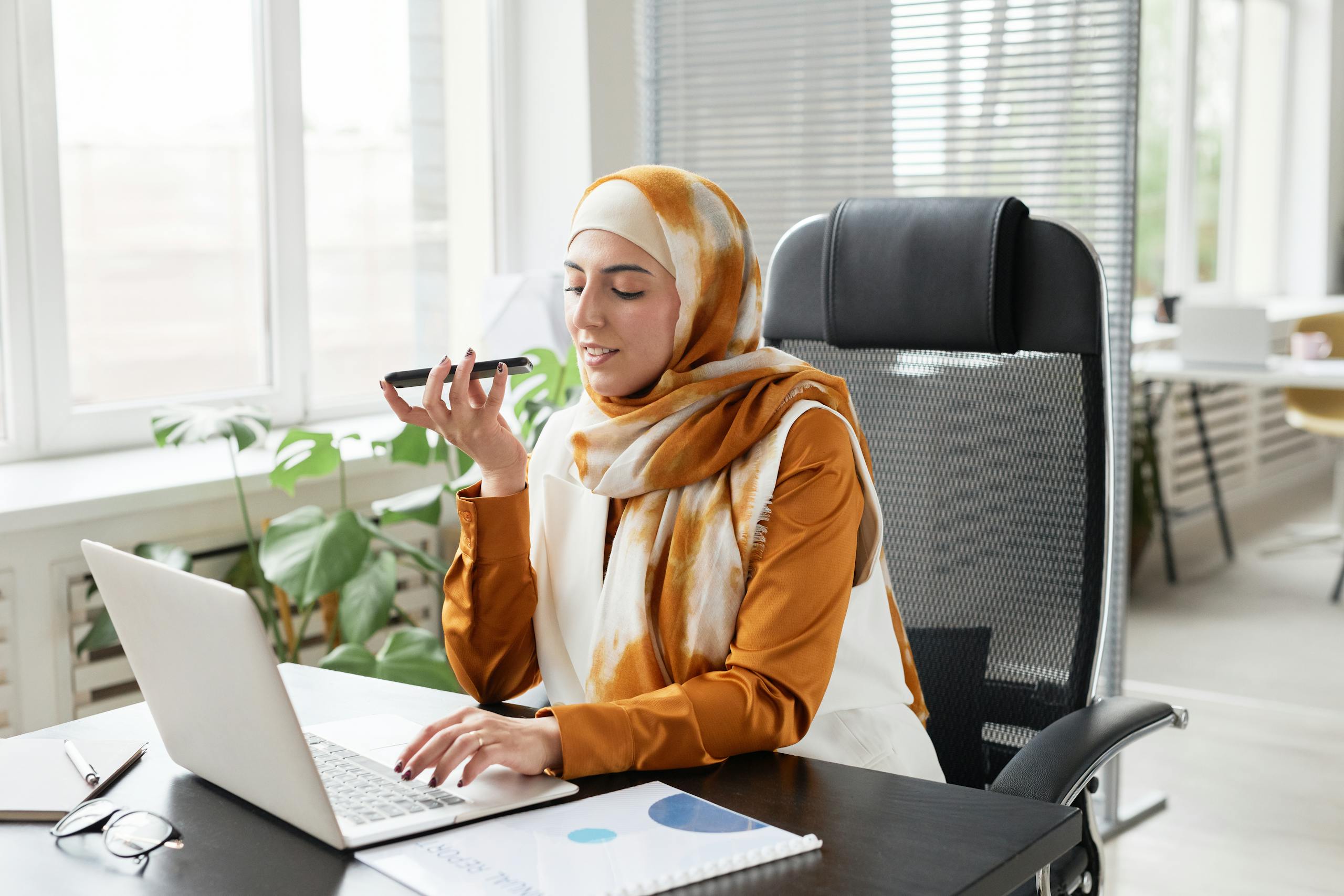 Muslim businesswoman in hijab working in a modern office, using a smartphone and laptop.