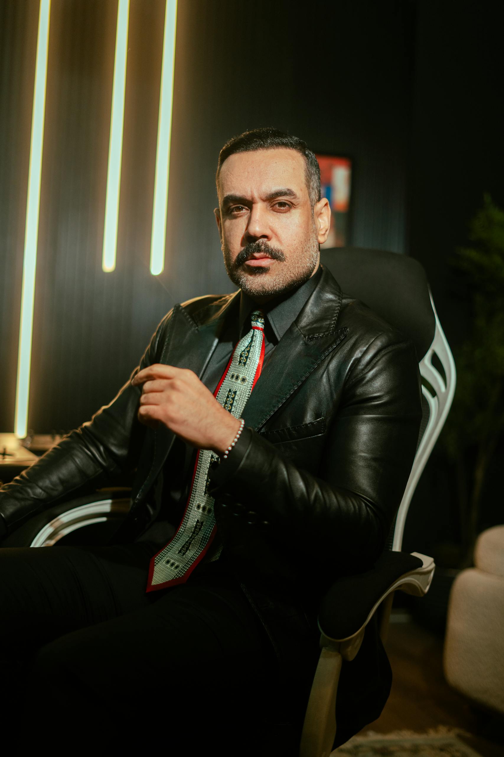 Portrait of a stylish man in a leather jacket sitting indoors with neon lights in Baghdad, Iraq.
