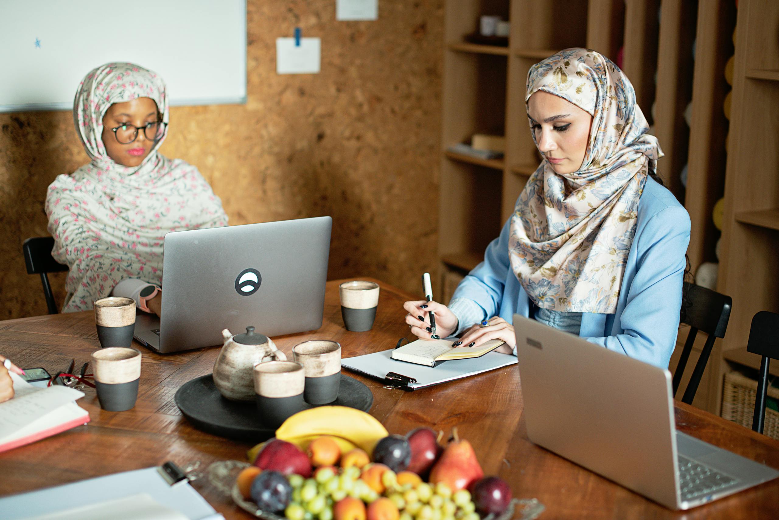 Two Muslim women in hijabs working together with laptops in a modern office setting.